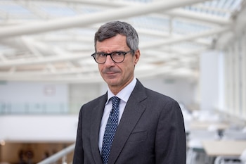 Retrato de un hombre de cabello cano con gafas, traje oscuro y corbata azul en un espacio interior bien iluminado