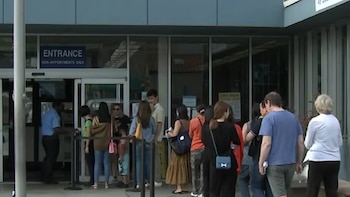 Varias personas de diversas edades hacen fila al aire libre frente a la entrada de un edificio con puertas de cristal y un letrero que indica 'ENTRANCE NON-APPOINTMENTS ONLY'