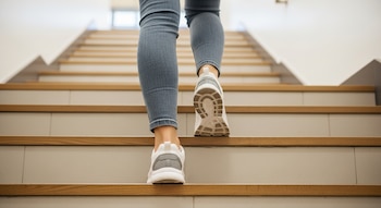 Un par de piernas con vaqueros azules y zapatillas blancas y grises suben una escalera de cemento y madera clara dentro de un edificio.
