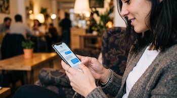 Una mujer de perfil sostiene un teléfono móvil con una conversación de chat en pantalla dentro de un café.