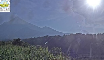Vista de montañas volcánicas, una con columna de humo. Vegetación densa en primer plano. Cielo azul con nubes y sol brillante. Logotipo del Instituto Nacional de Vulcanología