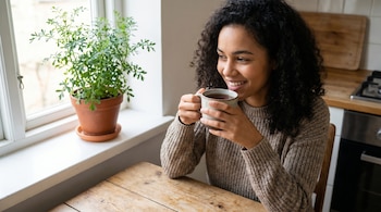 Mujer joven de cabello rizado y suéter marrón, sosteniendo una taza de té, sentada en mesa de madera junto a ventana con una planta de ruda en maceta.