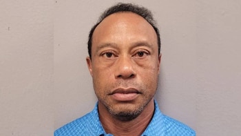 Primer plano frontal de Tiger Woods, un hombre con piel oscura y cabello oscuro, mirando directamente a la cámara, con una camisa azul estampada sobre un fondo liso