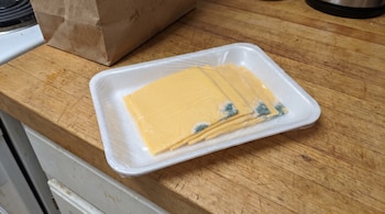 Bandeja de telgopor con rebanadas de queso amarillo y moho verde azulado, envuelta en plástico. Al fondo, una bolsa de papel y una superficie de madera.