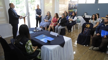 Un hombre en traje habla desde un atril a un grupo de personas sentadas en un salón. Una mesa cubierta con documentos y carpetas está en el primer plano