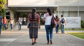 Vista trasera de dos mujeres jóvenes caminando en un campus universitario, una con traje indígena guatemalteco y otra con blusa bordada, jeans y mochila.