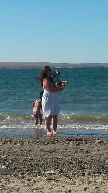 Una mujer con cabello oscuro y vestido blanco sostiene a un bebé, con una niña jugando en el agua detrás, y un paisaje de playa con mar y cerros al fondo
