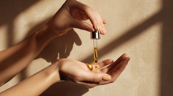 Dos manos, un gotero de vidrio con líquido dorado, gotas de líquido dorado en una palma, fondo texturizado de color beige con sombras.