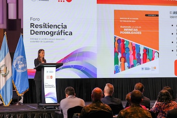 Una mujer habla en un podio. Al lado, banderas de Guatemala y Naciones Unidas. Detrás, una pantalla muestra el Foro Resiliencia Demográfica y un informe de población 2024