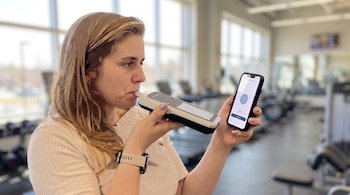 Mujer rubia con camisa clara sostiene un dispositivo sensor de aliento junto a su boca y un teléfono inteligente con una aplicación abierta, en un gimnasio.