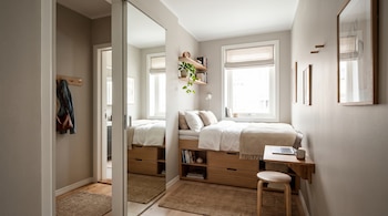 Una cama elevada sobre estantes de madera, un espejo alto, un escritorio pequeño con un ordenador portátil, un taburete y una ventana al fondo.