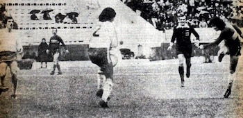 Cancha de fútbol con cinco jugadores de dos equipos, uno con camiseta número 7, y un árbitro. Las tribunas de un estadio al fondo están ocupadas por espectadores