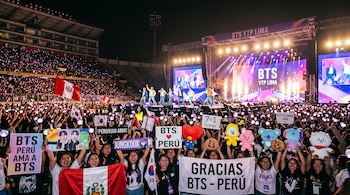 Multitud en un estadio sostiene pancartas, banderas de Perú y Corea del Sur, y peluches frente a un escenario con pantallas y luces.