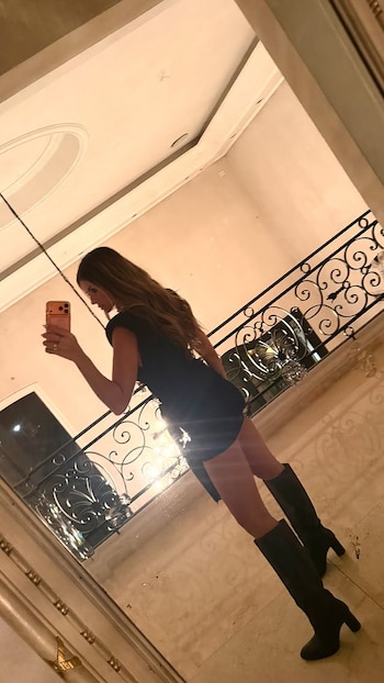 Mujer de cabello largo con vestido y botas negras, tomando una selfi con un teléfono en un espejo, junto a una barandilla de hierro forjado