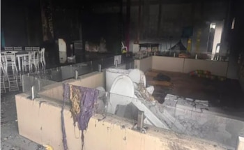 Interior de un local con paredes ennegrecidas, techo y mobiliario calcinados. Mesas, sillas y escombros esparcidos en el suelo con restos de agua