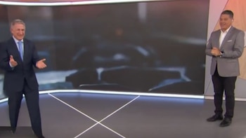 Dos hombres con trajes, uno azul y otro gris, ríen de pie en un estudio de televisión moderno con una pantalla grande al fondo