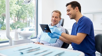 Un dentista con guantes azules sonríe mientras muestra una tableta a un paciente sentado en una silla dental. Se ven instrumentos odontológicos en primer plano.