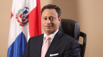 Hombre sonriente con traje oscuro, camisa blanca y corbata rosa sentado frente a una bandera de la República Dominicana y una pared beige.
