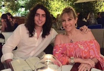 Un hombre de cabello largo ondulado y una mujer rubia con vestido estampado rosa sonríen sentados frente a una vela en una mesa