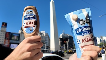 Dos manos sostienen productos Hellmann's sabor asado: una botella y un envase flexible, con el Obelisco de Buenos Aires al fondo bajo un cielo azul claro