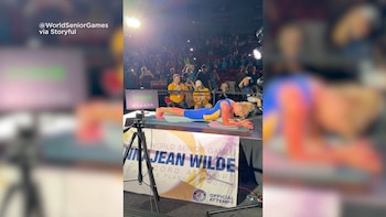 Una mujer realiza una plancha sobre una colchoneta en un escenario frente a un público, junto a un cronómetro digital y una pancarta