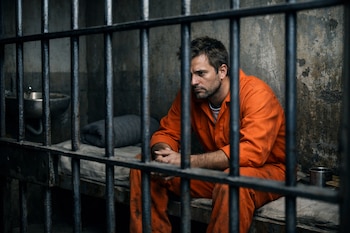 Un hombre de mediana edad con uniforme naranja está sentado en una cama dentro de una celda de prisión, vista a través de los barrotes.