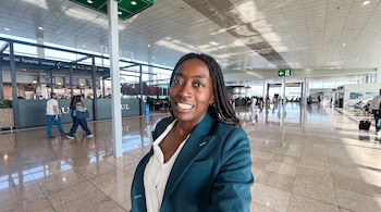 Una mujer sonríe en primer plano dentro de una terminal de aeropuerto con pisos pulidos, locales y viajeros al fondo.
