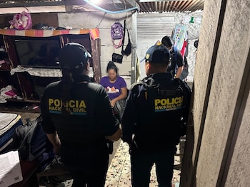 Dos agentes con uniformes de Policía Nacional Civil de espaldas, una mujer sentada, y un hombre de pie dentro de una vivienda con mobiliario diverso