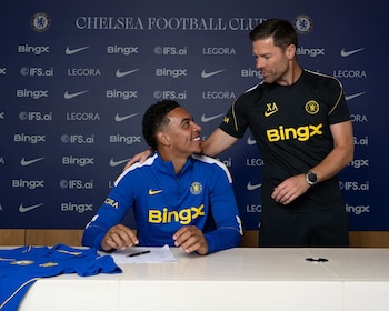 Jugador con camiseta azul sentado firmando. Hombre con camiseta negra de pie a su lado. Fondo azul con escudo Chelsea F.C. y logos de Bingx, IFS.ai, Legora, Nike