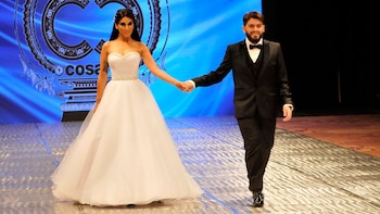Diego Maradona JR, con esmoquin negro, y Nunzia Pennino, con vestido de novia blanco, caminan de la mano sobre una pasarela brillante con un fondo azul