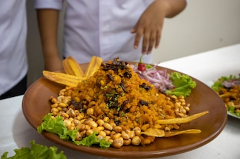 Plato marrón con montaña de Seco de Chabelo, maíz cancha, chifles. Guarnición de lechuga, cebolla roja, ají. Dos personas en segundo plano