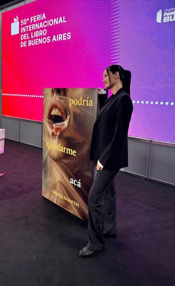 Oriana Sabatini, con blazer y pantalón negro, posa sonriente junto a una réplica gigante de la portada de su libro. Al fondo, pantalla con logo de la 50° Feria del Libro