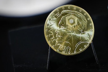 Moneda de oro redonda con el perfil estilizado de una figura femenina geométrica en el anverso, sobre un fondo oscuro y borroso, sostenida por un soporte claro