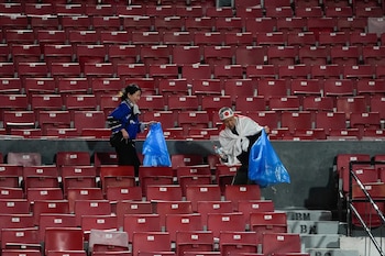 Dos aficionados japoneses, uno con una diadema con bandera japonesa, recogen basura en bolsas azules entre las gradas rojas vacías de un estadio