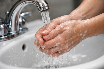 Un grifo de metal plateado vierte agua sobre unas manos con espuma en un lavamanos blanco.