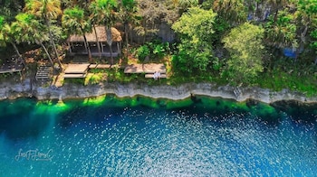 Vista aérea de un cuerpo de agua azul y verde, orilla rocosa, vegetación abundante, palmeras, un quiosco de techo de paja, plataformas de madera, escaleras y dos personas