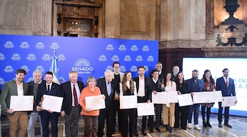 Grupo de dieciocho personas con diplomas y trofeos ante un telón azul con logos "Senado Argentina", una bandera y columnas de mármol.