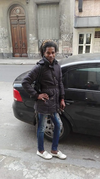 Una persona con rastas, chaqueta negra, vaqueros rotos y zapatillas blancas, de pie junto a un coche negro en una calle urbana con edificios