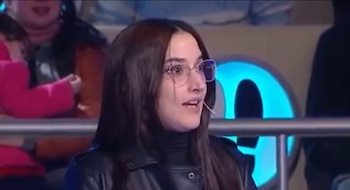 Mujer joven con cabello oscuro, anteojos, chaqueta negra, micrófono en el cuello. Habla en un set con un objeto azul luminoso y público borroso