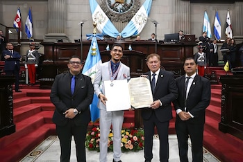 Cuatro hombres posan en un salón de eventos oficial. Uno sonríe con una medalla y un diploma, otro sostiene un documento. Banderas y público de fondo