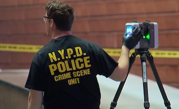 Oficial de la NYPD con camiseta "CRIME SCENE UNIT" opera un escáner 3D en una escena del crimen, con cinta amarilla de seguridad de fondo