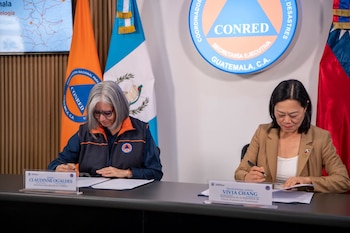 Dos mujeres sentadas en una mesa, firmando documentos. Una bandera de Guatemala y una bandera naranja de CONRED. Logo de CONRED en la pared