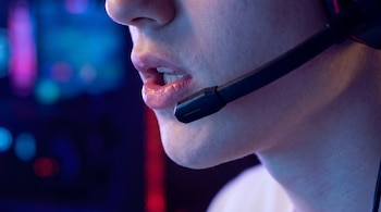 Primer plano de la boca de una persona usando un micrófono de casco gamer. Los labios están entreabiertos, con iluminación azul y roja de fondo desenfocado.