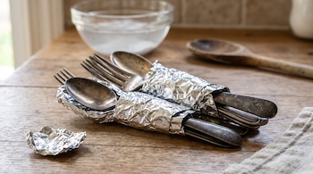 Cubiertos de metal envueltos en papel de aluminio sobre una mesa de madera, junto a un tazón de vidrio y una cuchara de madera.