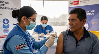 Un hombre recibe una inyección en el brazo de una mujer con mascarilla, bata azul y guantes, frente a otra persona con mascarilla y un cartel de MINSA.