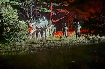 Vehículo militar camuflado con faros traseros encendidos y varios soldados con uniformes de camuflaje en un bosque oscuro, bajo árboles altos