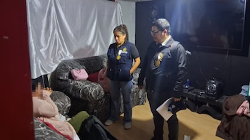 Dos agentes de la Fiscalía, un hombre y una mujer con chalecos oficiales, inspeccionan una sala. Se ven sofás, cortinas y una persona borrosa