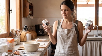 Una mujer con delantal en una cocina, sostiene un frasco de polvo para hornear en la mano izquierda y un frasco de bicarbonato de sodio en la derecha.