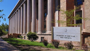 Edificio imponente del Poder Judicial de Mendoza con columnas clásicas, un cartel con el nombre y áreas verdes con césped y arbustos bajo un cielo azul