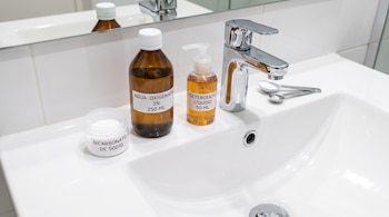 Tres recipientes con etiquetas de bicarbonato, agua oxigenada y detergente líquido junto a un grifo metálico y un lavabo de cerámica blanca.